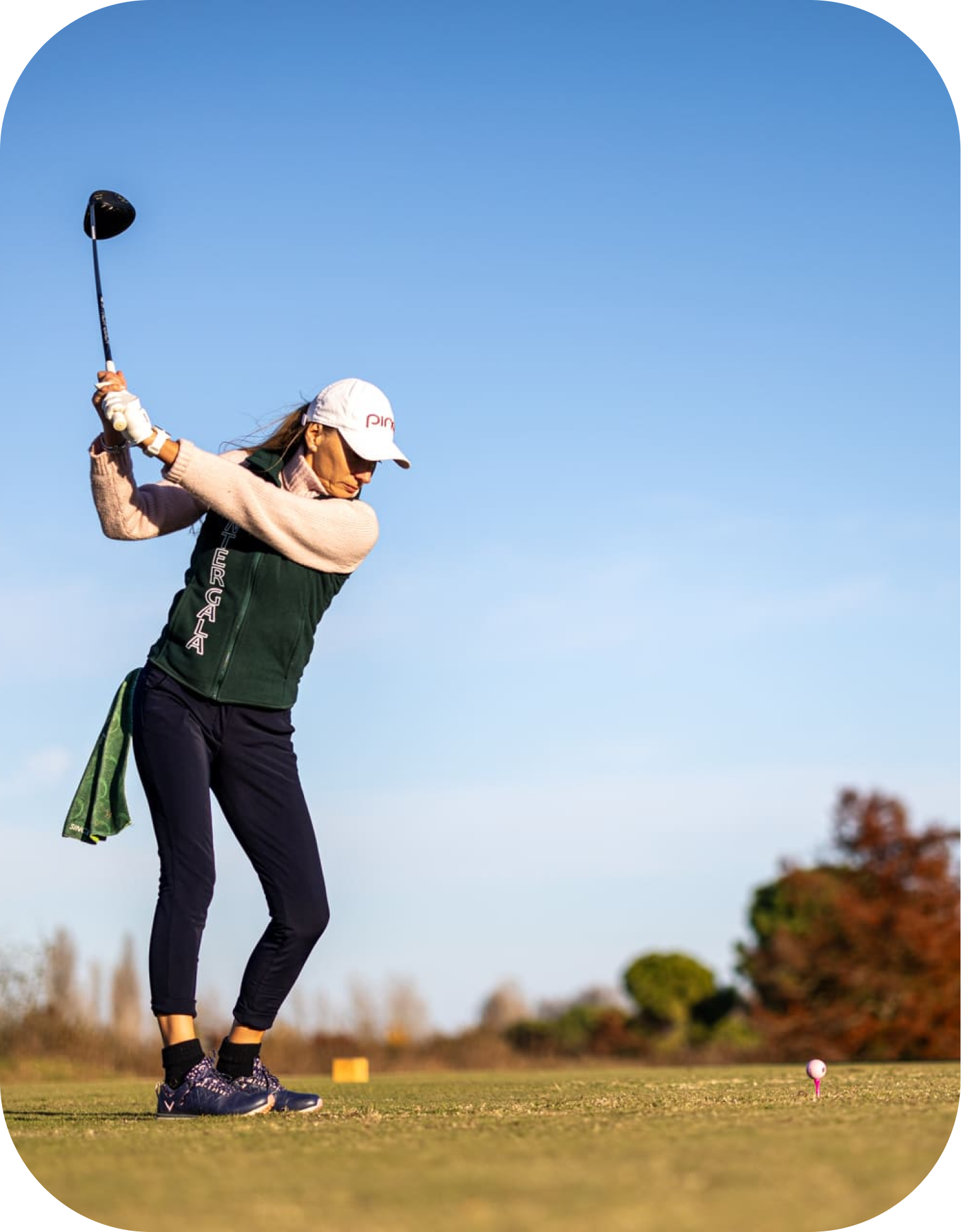 donna-golf Golfista donna in azione sul tee di partenza, intenta a completare il backswing con un driver sotto un cielo sereno. Indossa un cappellino bianco e un gilet verde, con sullo sfondo alberi autunnali.